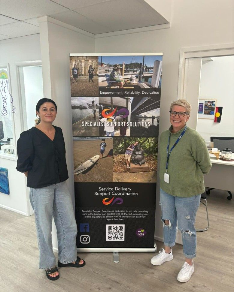 Two Women Are Standing Next to Each Other in Front of a Banner — Specialist Support Solutions in Yeppoon, QLD