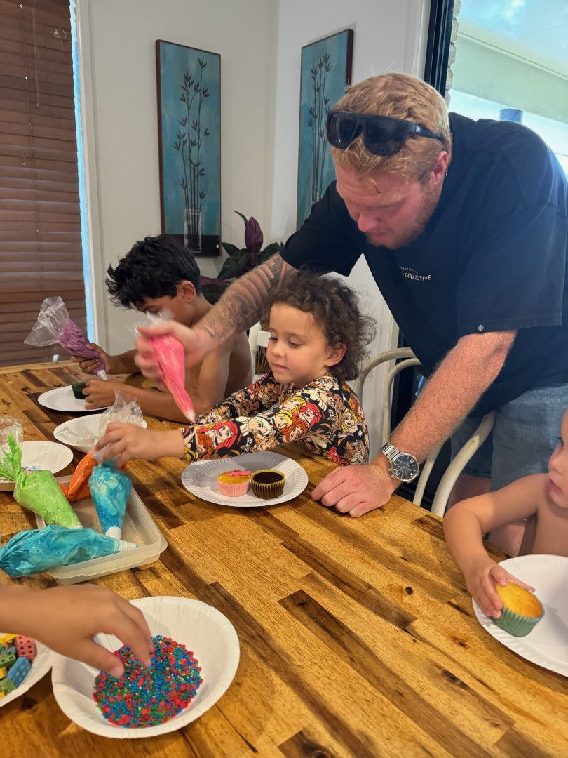 A person assists a child in decorating cupcakes with colored icing at a wooden table, while others join in the activity.— Specialist Support Solutions in Rockhampton City, QLD