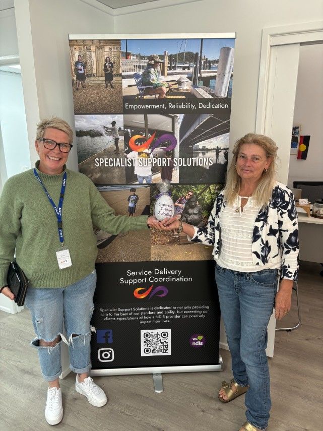 Two Women Are Standing Next to Each Other in Front of a Banner — Specialist Support Solutions in Emerald, QLD