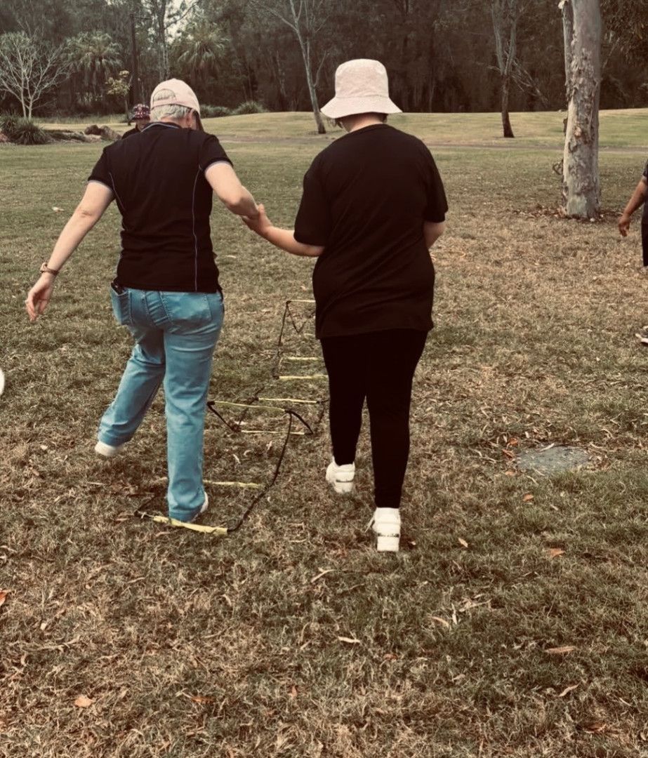 Two people wearing black shirts and hats hold hands while walking through an agility ladder set up in a grassy field — Specialist Support Solutions in Rockhampton City, QLD
