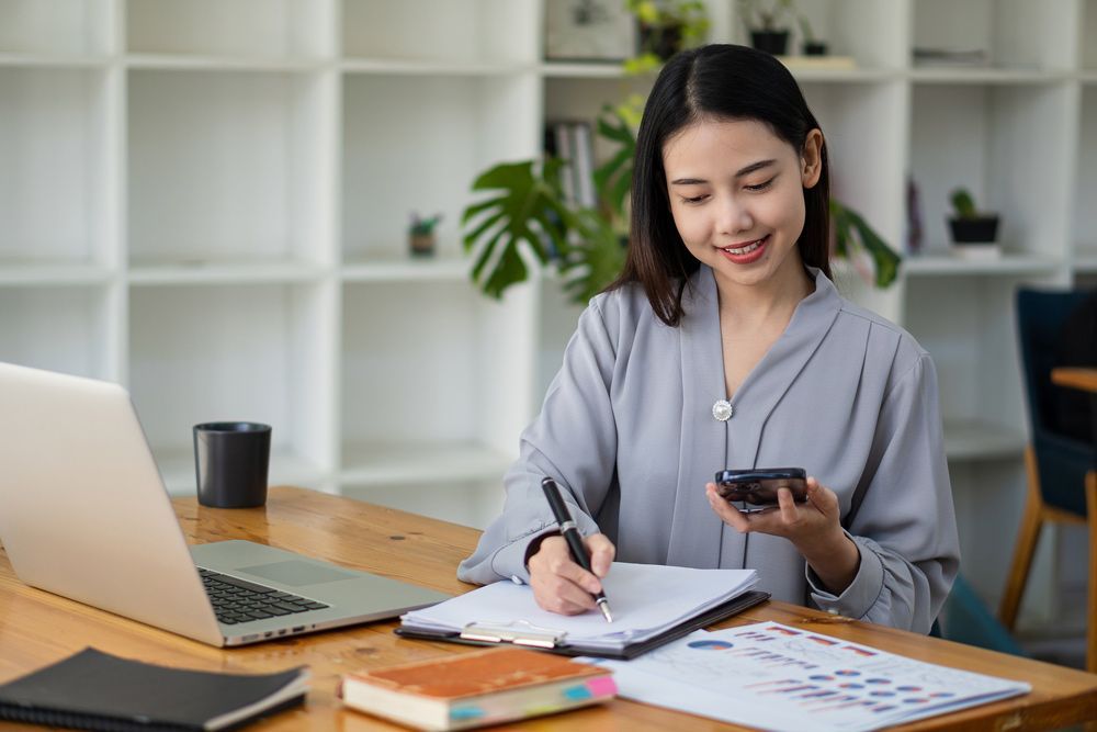 A Woman Is Sitting At A Desk With A Laptop And A Cell Phone — Bosco Accounting Pty Ltd In Warrawong, NSW