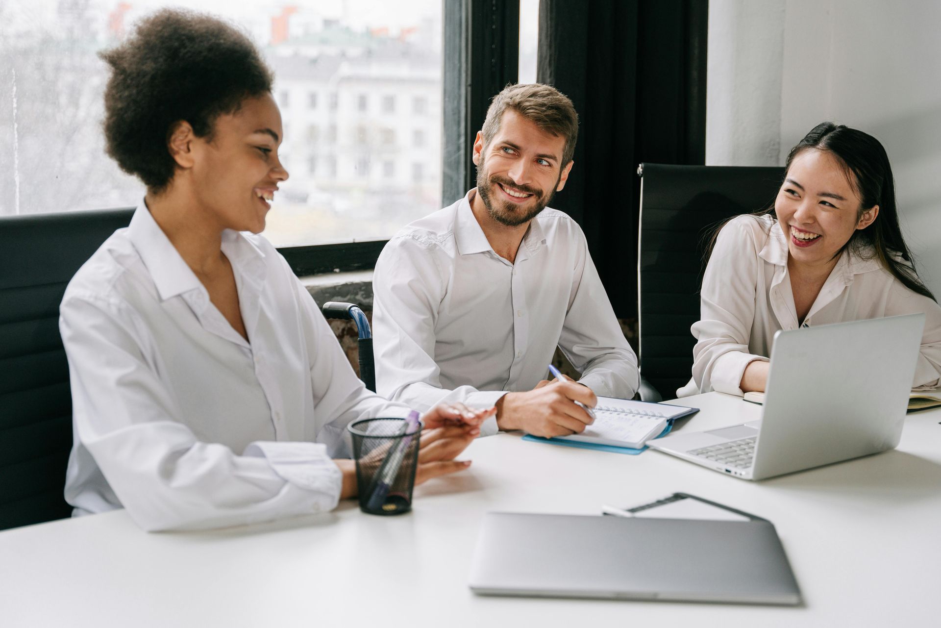 Three people smiling around a table with a laptop, notebooks, and a pen holder in an office setting — Bosco Accounting Pty Ltd In Warrawong, NSW