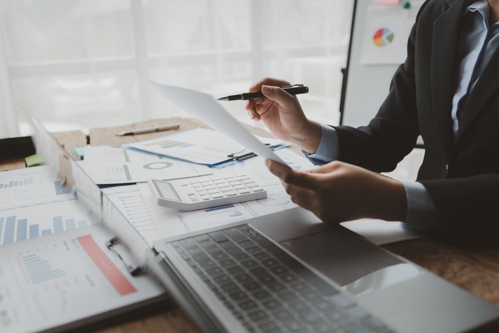 Person in a suit reviews financial documents, pointing with a pen, surrounded by charts and a calculator at a desk — Bosco Accounting Pty Ltd In Warrawong, NSW