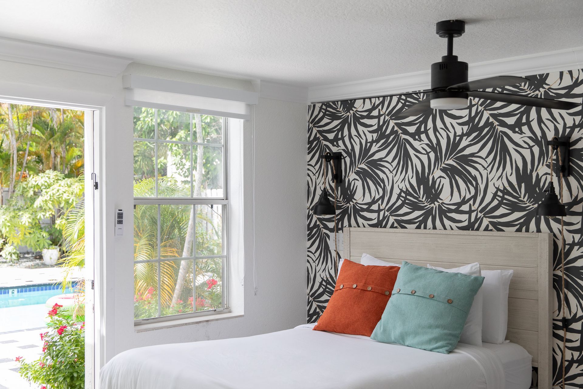 Bedroom with bed, patterned wallpaper, pool view, window, white walls, and a ceiling fan.