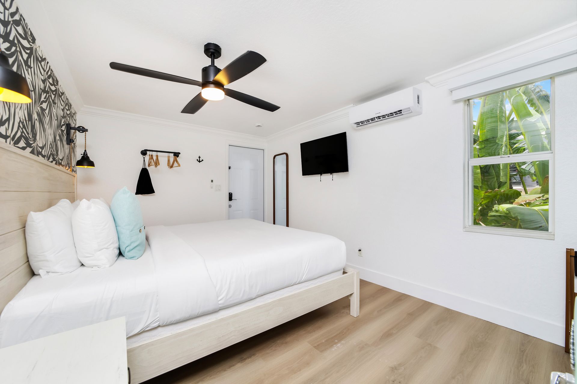 Bedroom with white walls, bed, TV, ceiling fan, and window overlooking greenery.