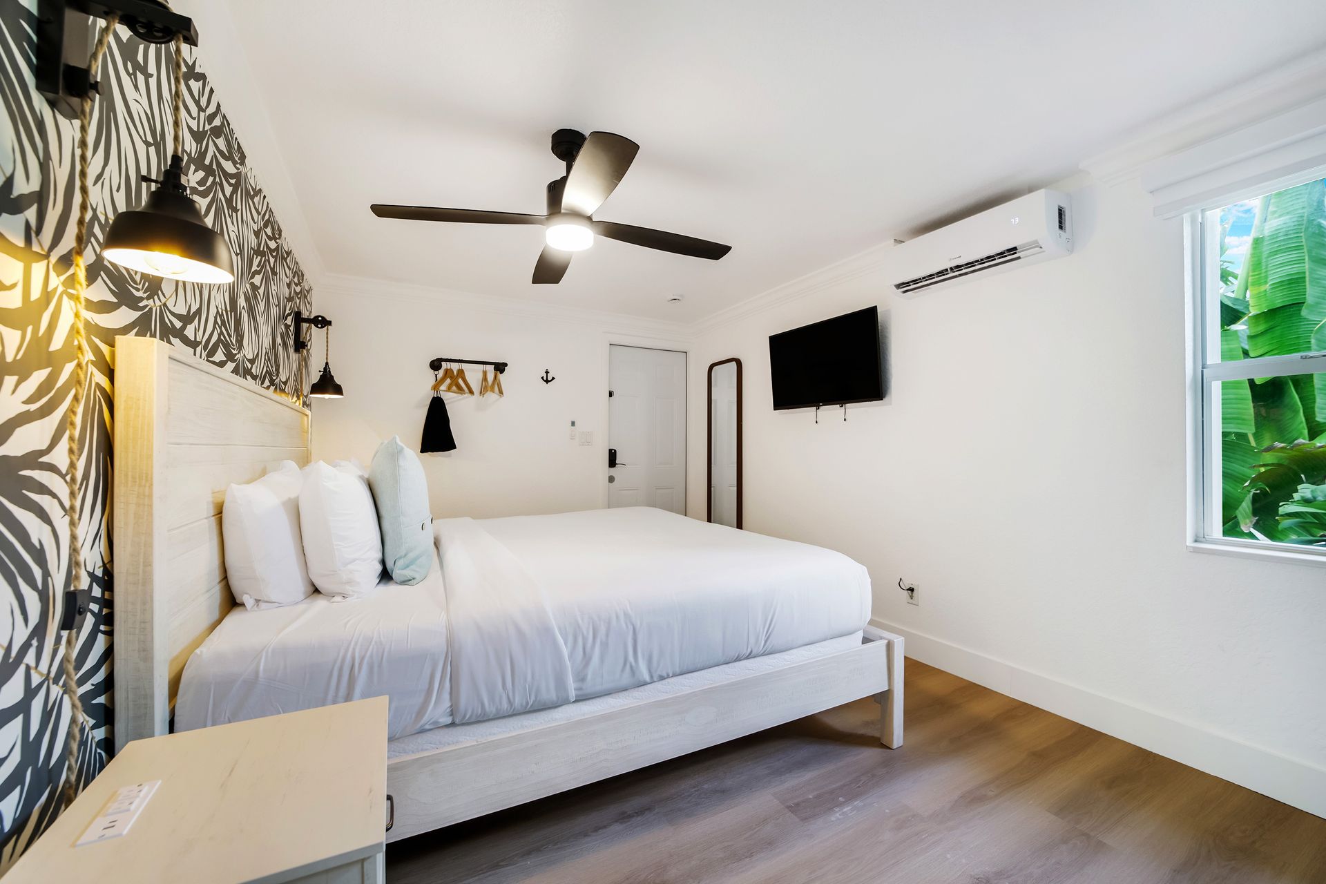 Bedroom with a white bed, patterned wallpaper, a TV, and a window with greenery.
