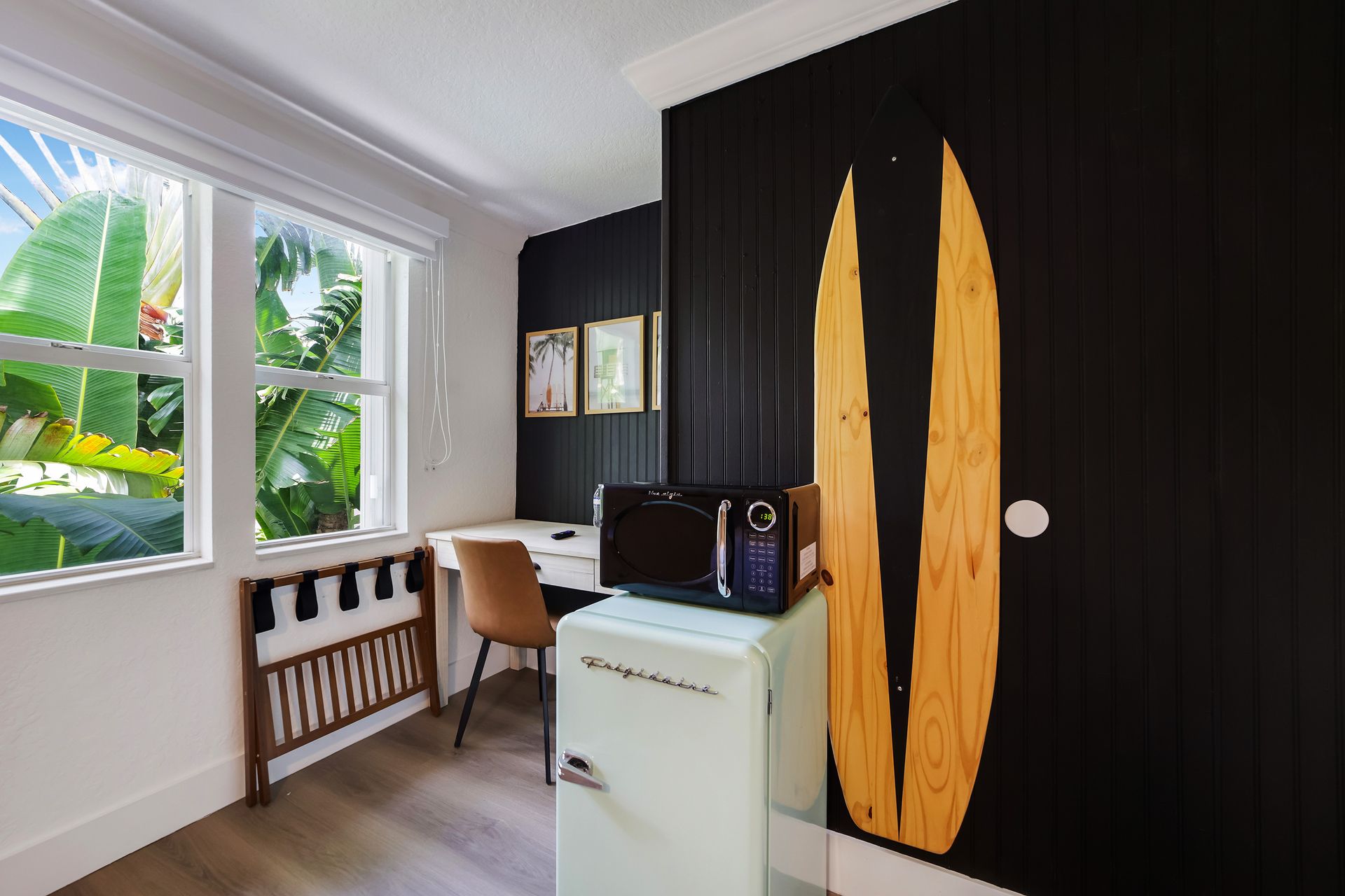 Small hotel room with black accent wall, surfboard art, mini-fridge, and desk with window view.