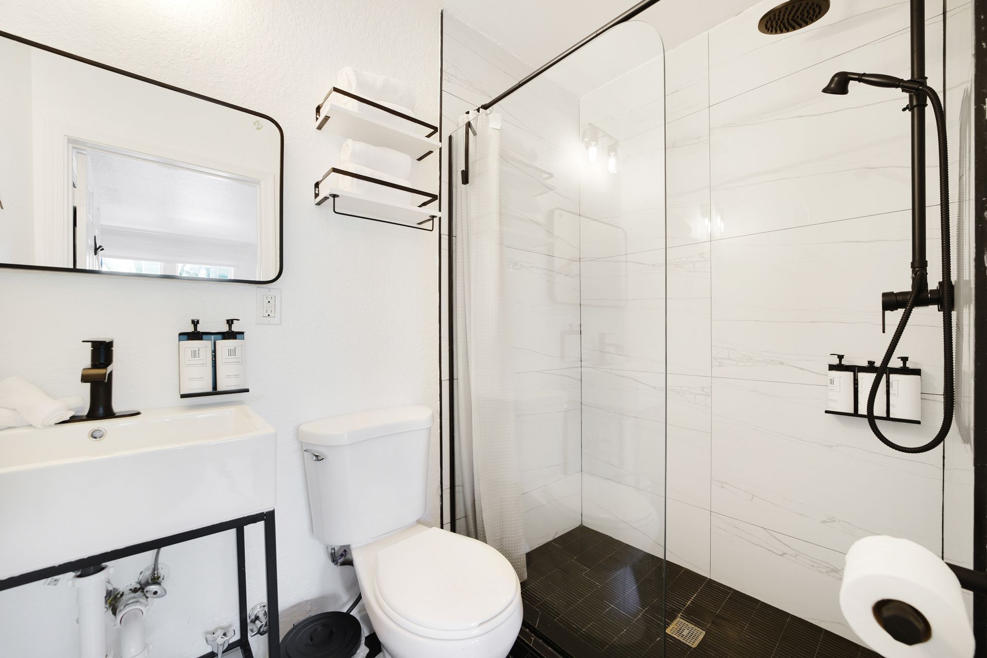 Modern white bathroom with black accents. Features a shower, sink, toilet, and mirror.