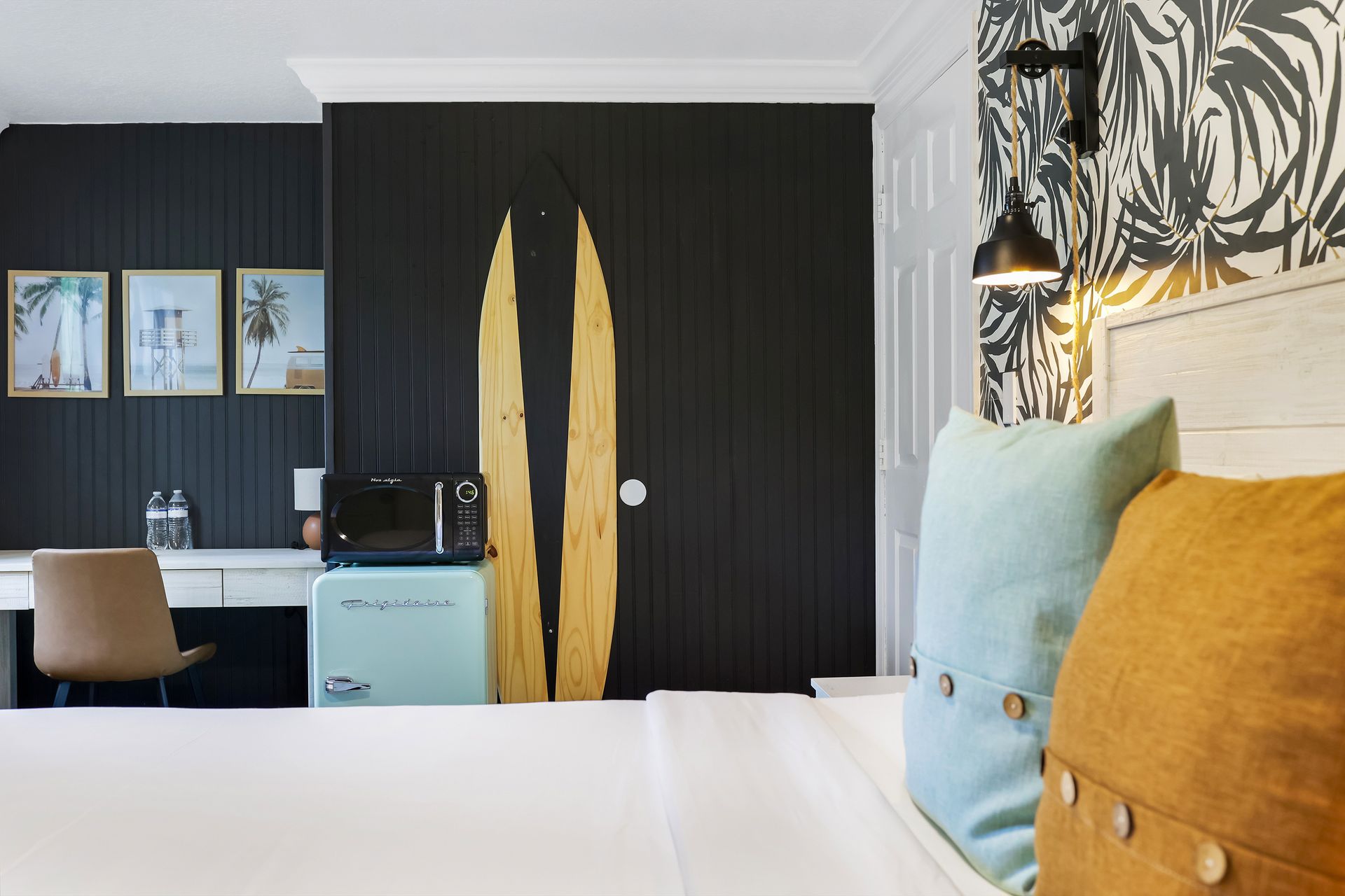Bedroom with bed, desk, black wall, decorative surfboards, mini-fridge, and framed art.