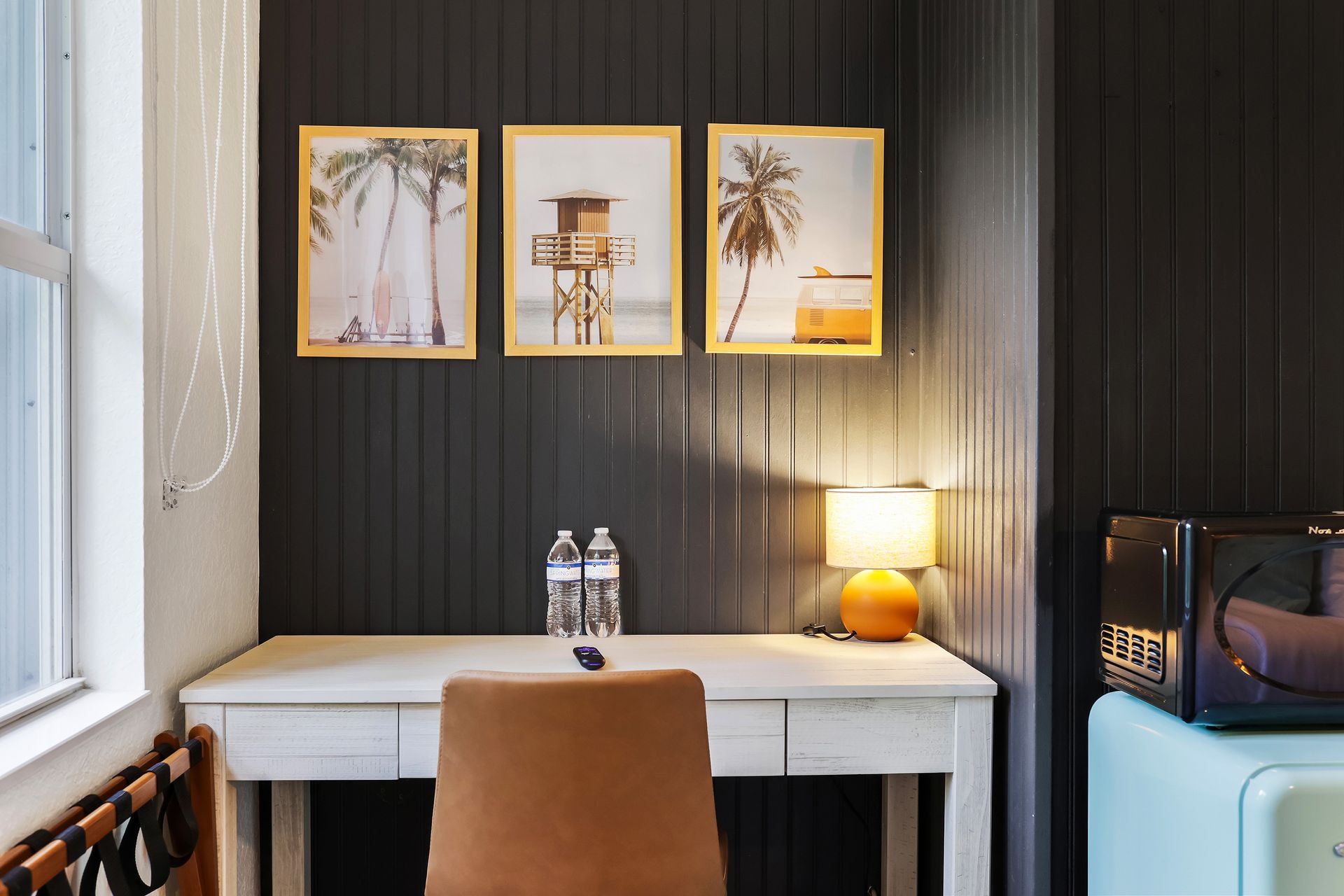 Desk with three framed beach prints, lamp, water bottles, chair, and mini-fridge against a dark paneled wall.