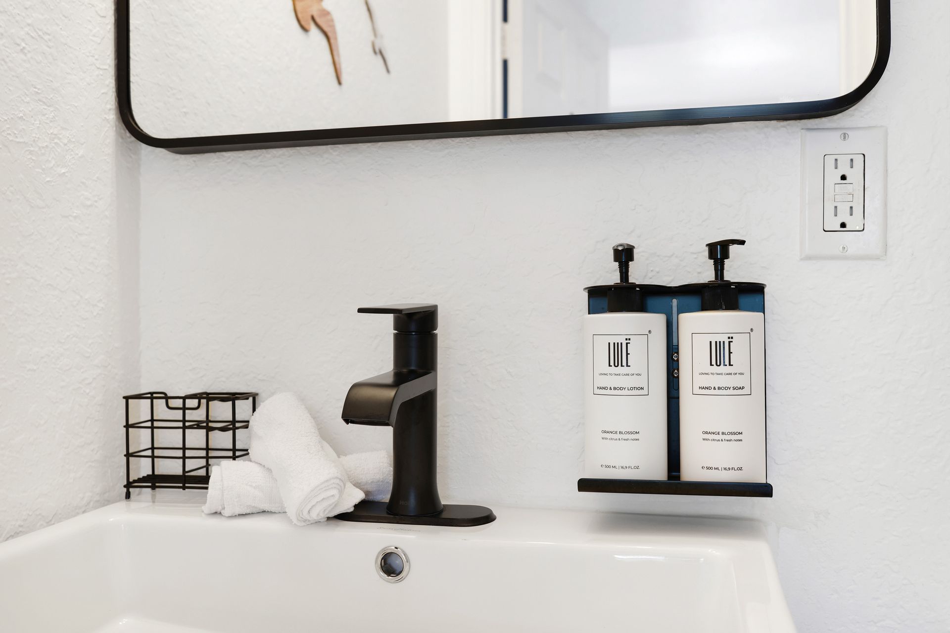 White bathroom sink with black faucet, soap dispensers, and small wire basket.