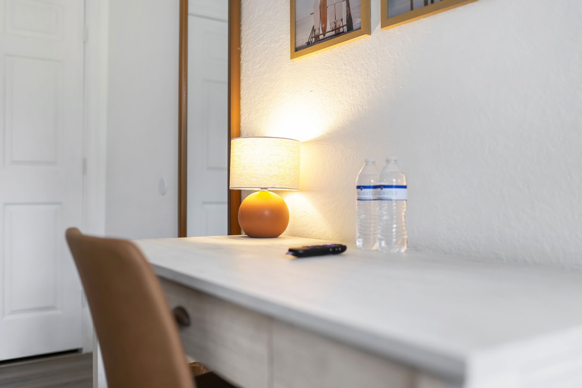 Desk with lamp, water bottle, and remote. Chair in front of a white wall with a door and mirror.