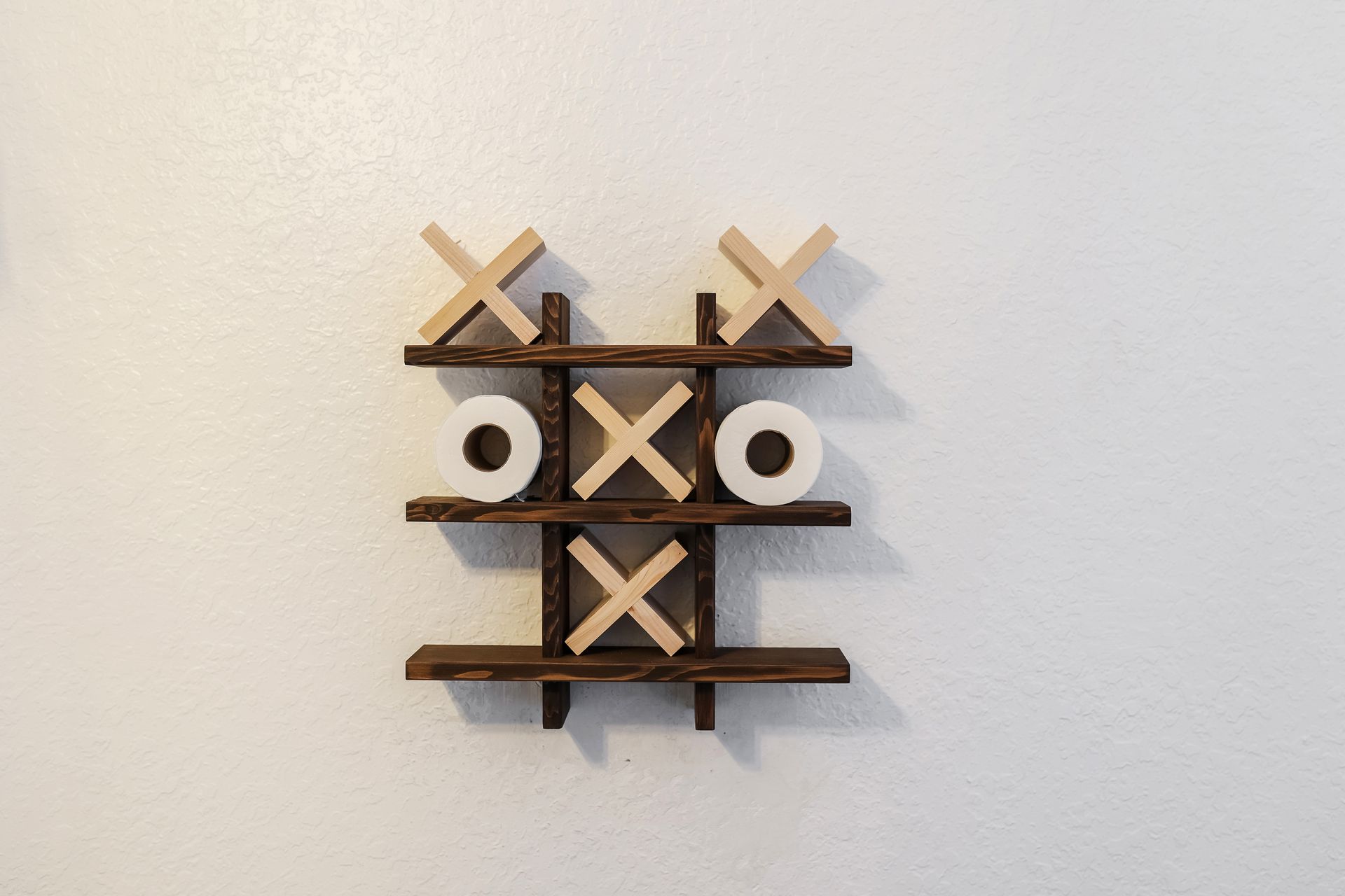 Tic-tac-toe game board made of wood; has three Xs and two Os, mounted on a white wall.