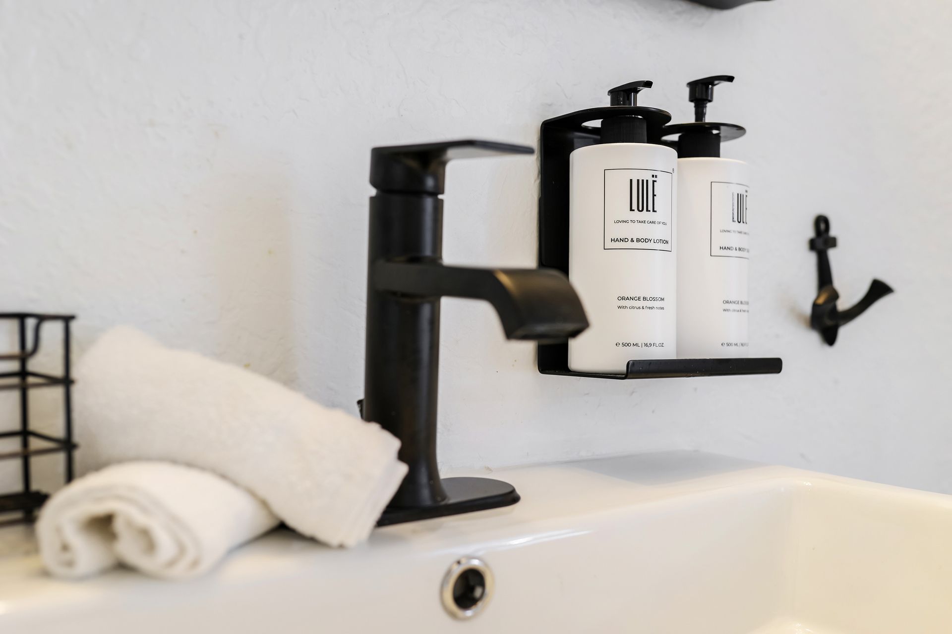 Black faucet and toiletries on a white sink, with towels and a shelf with soap bottles.