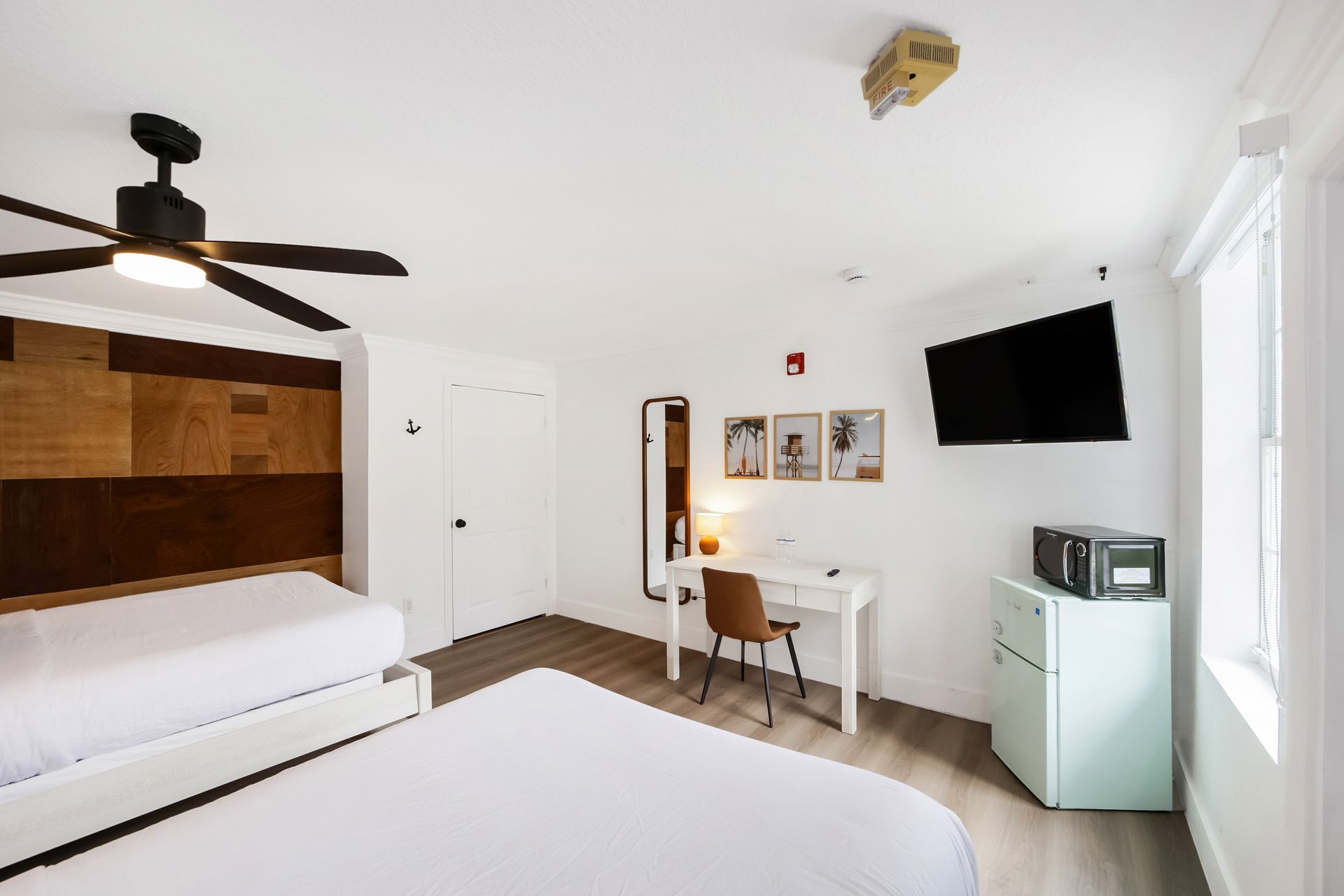 Hotel room with two beds, desk, TV, refrigerator, and ceiling fan. White and wood tones.
