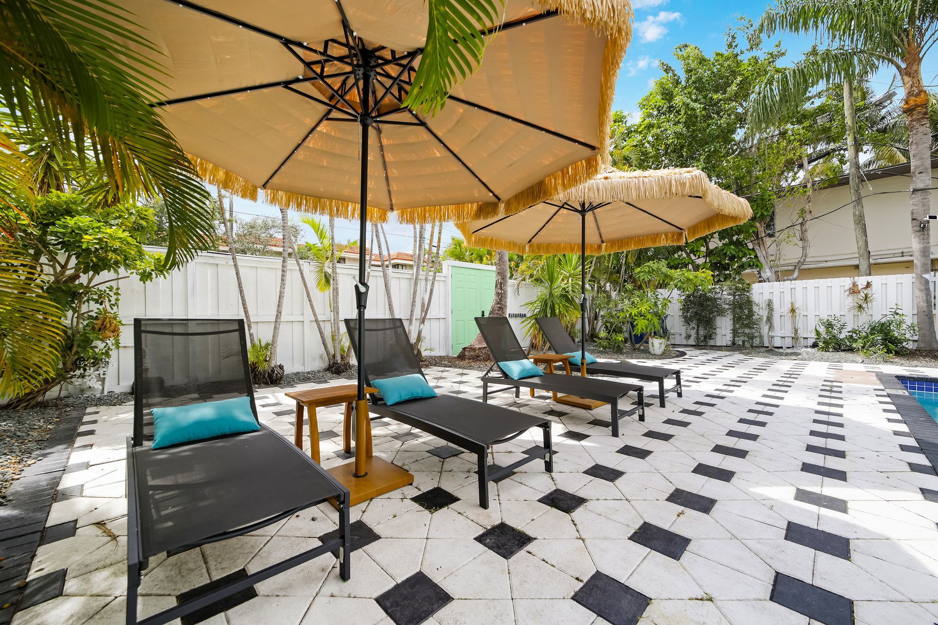 Lounge chairs under fringed umbrellas on a patterned patio next to greenery.