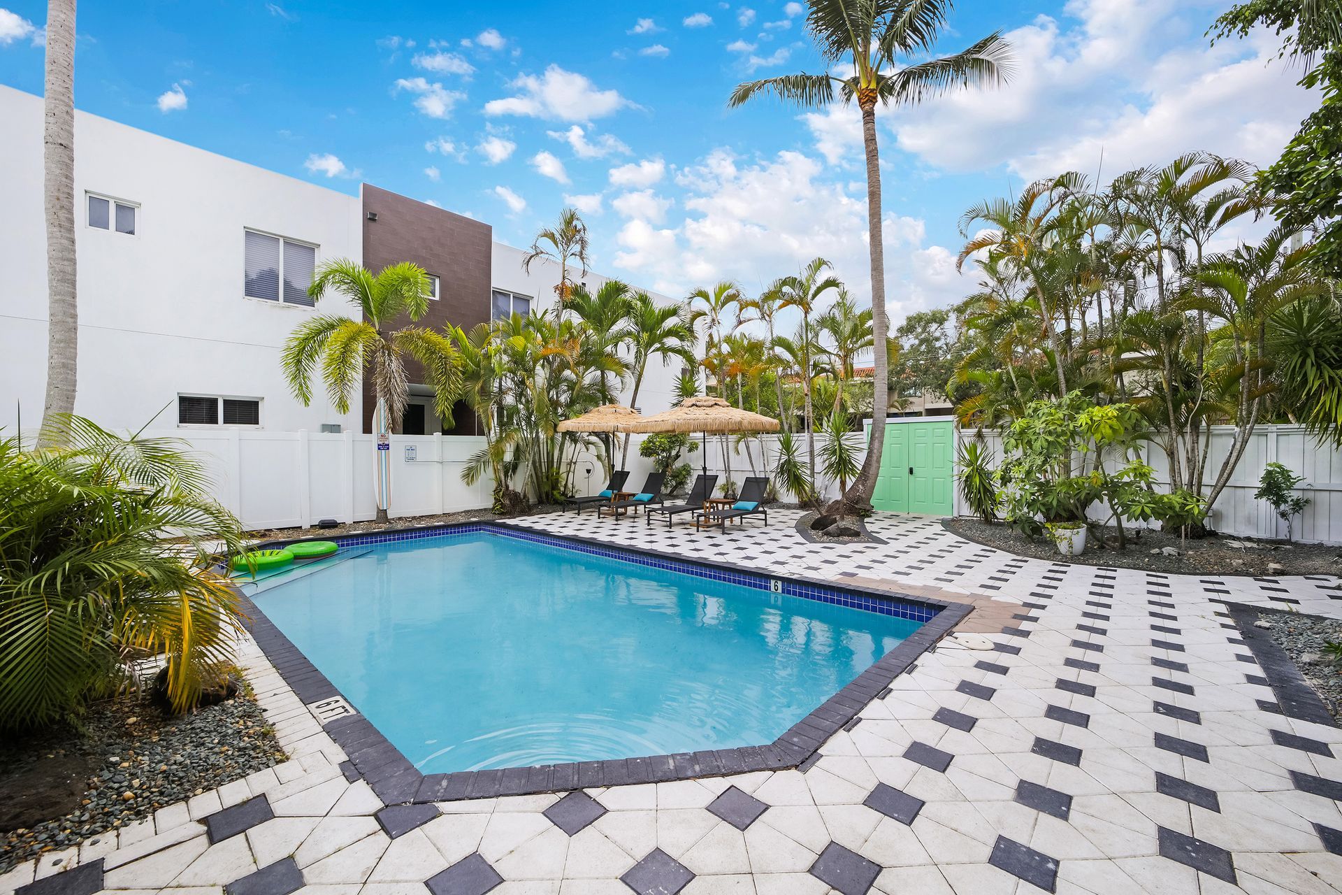 Swimming pool in a backyard with patio, tropical trees, and modern white building.