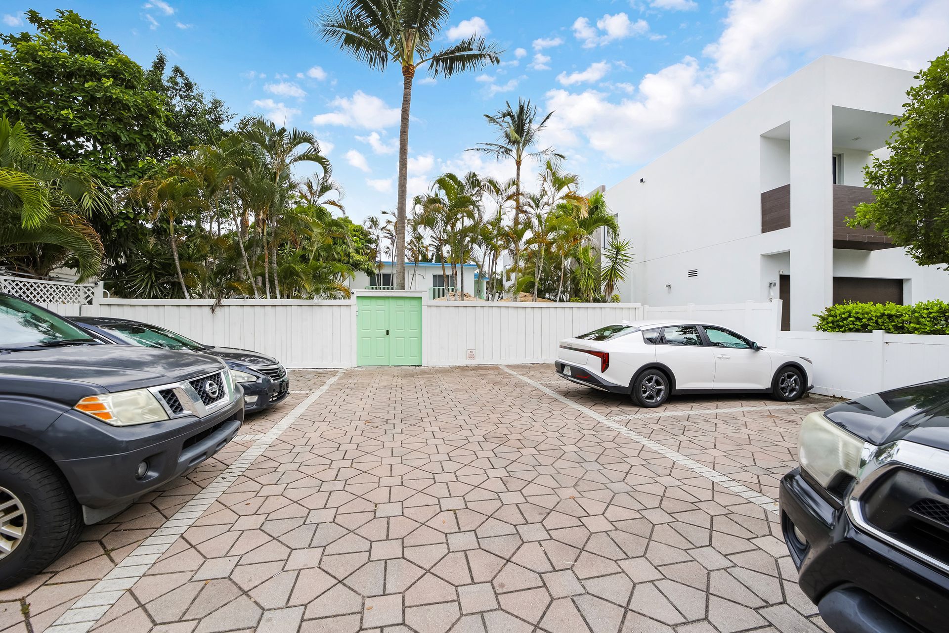 Parking lot with several cars parked on a textured stone surface, beside a white building and green trees.