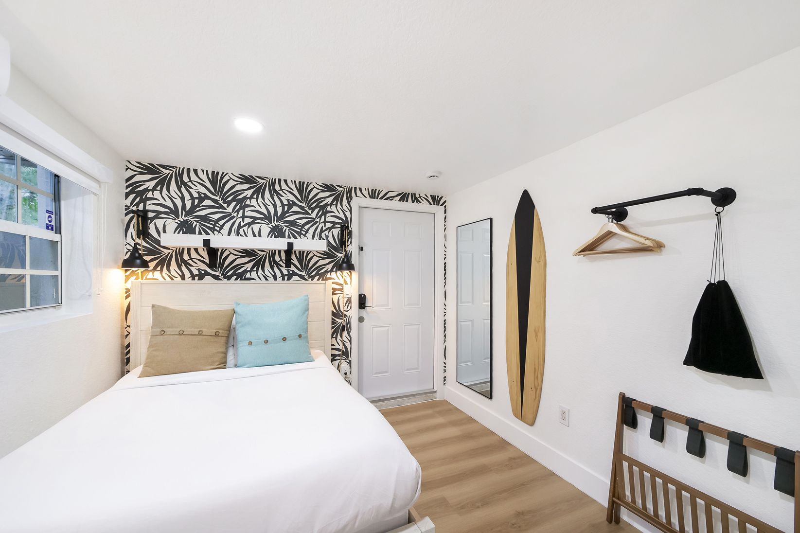 Bedroom with white walls, patterned accent wall, bed with pillows, surfboard, mirror, and coat rack.