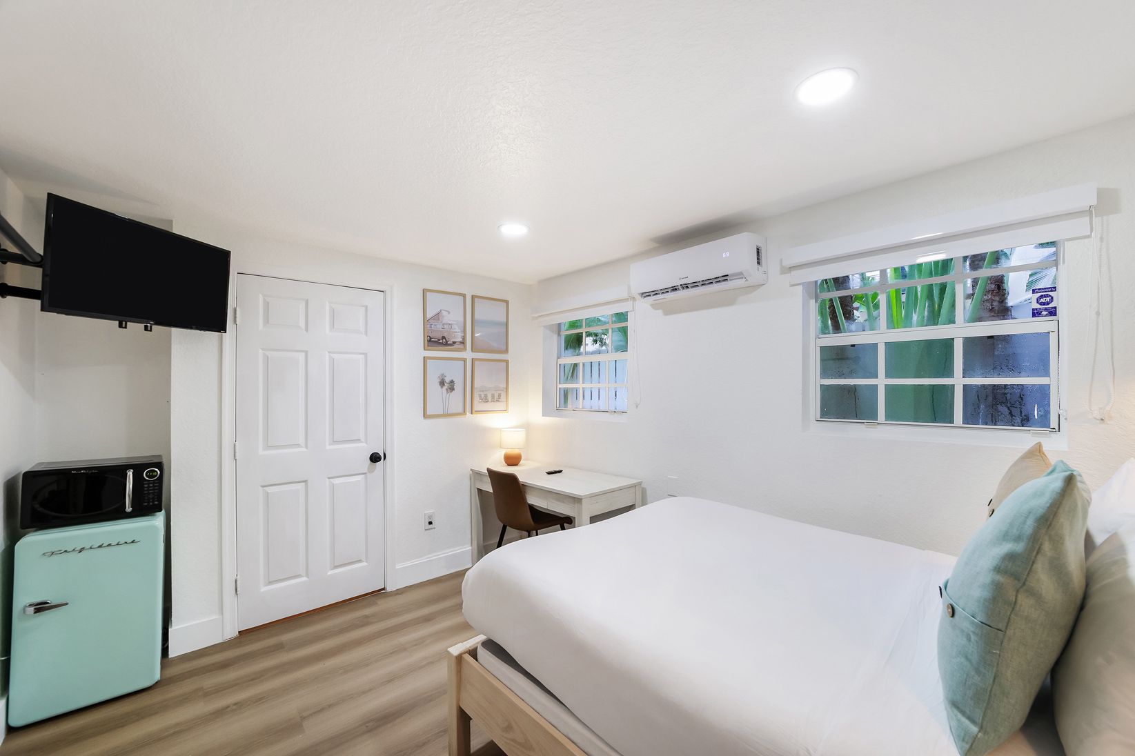 Bright white bedroom with bed, desk, wall-mounted TV, and a teal refrigerator.