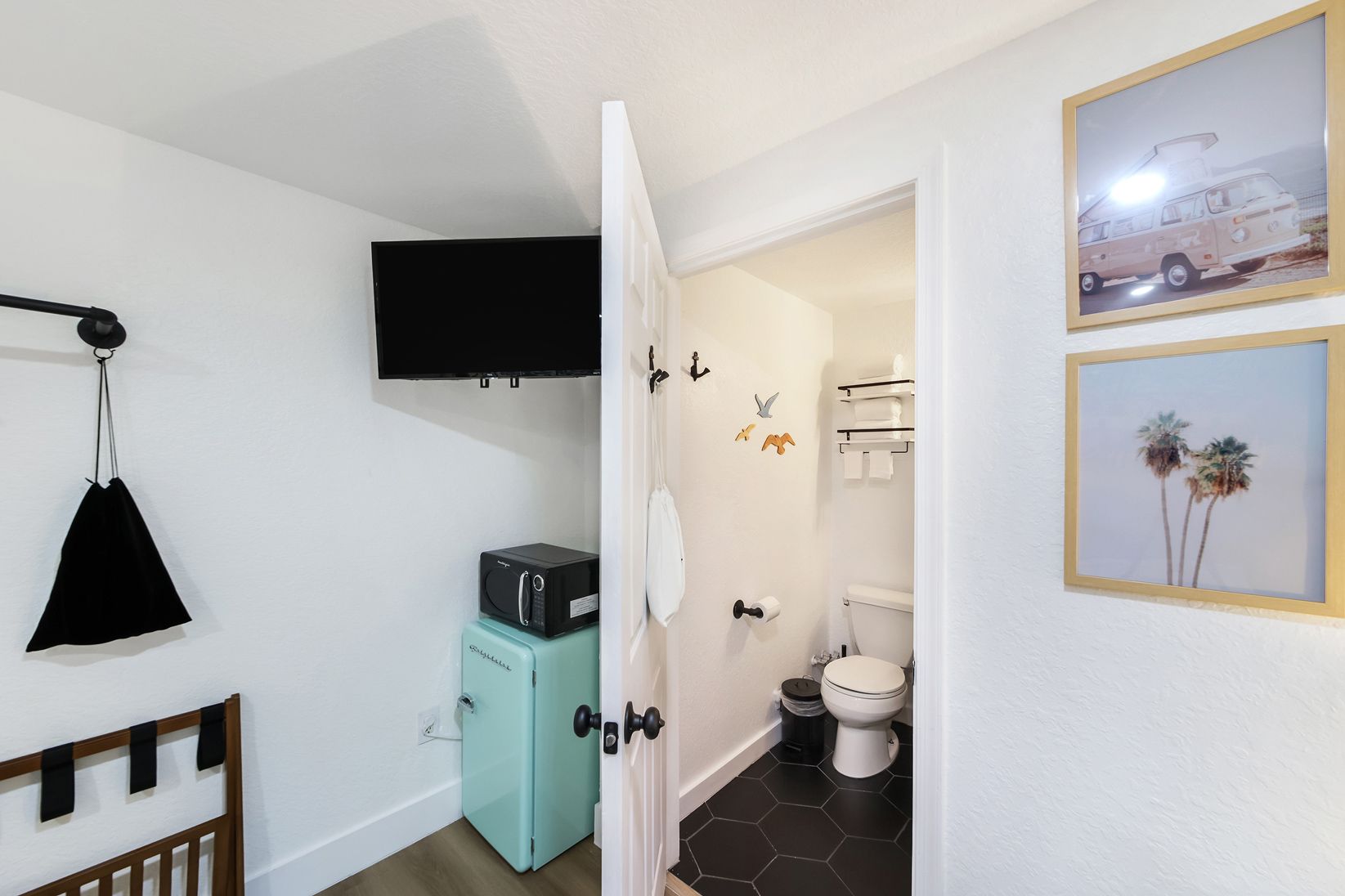 View into a small bathroom from a room; a refrigerator, TV, and framed art are visible.