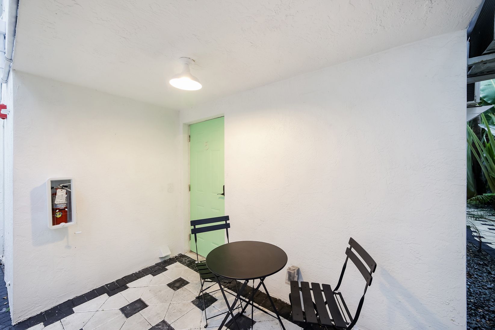 Small outdoor seating area: black table and chair, white walls, green door, tiled floor, fire extinguisher.
