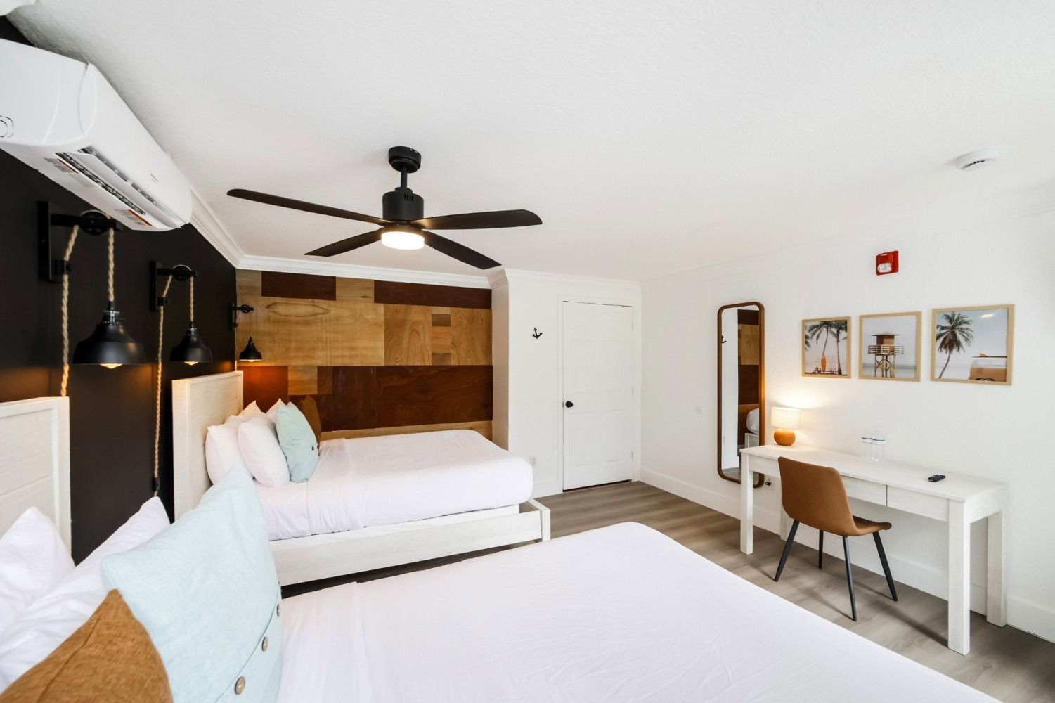 Hotel room with two beds, desk, mirror, and art. Neutral tones, bright.