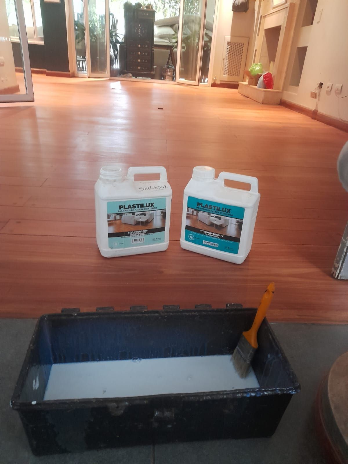 Dos recipientes para líquidos y una bandeja de pintura con pincel sobre un piso de madera.