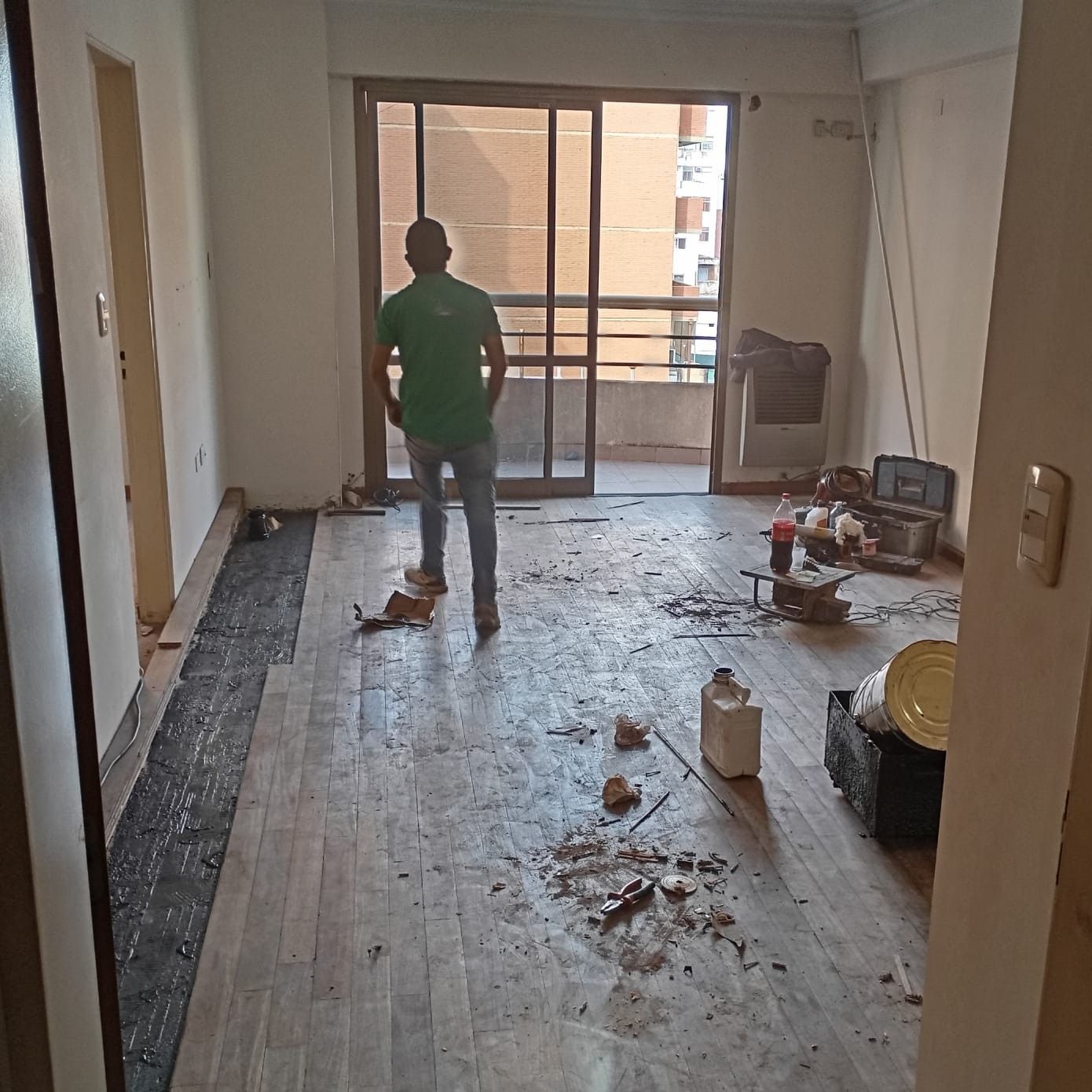 Un hombre con camisa verde se encuentra en una habitación con balcón, hay escombros en el piso de madera.