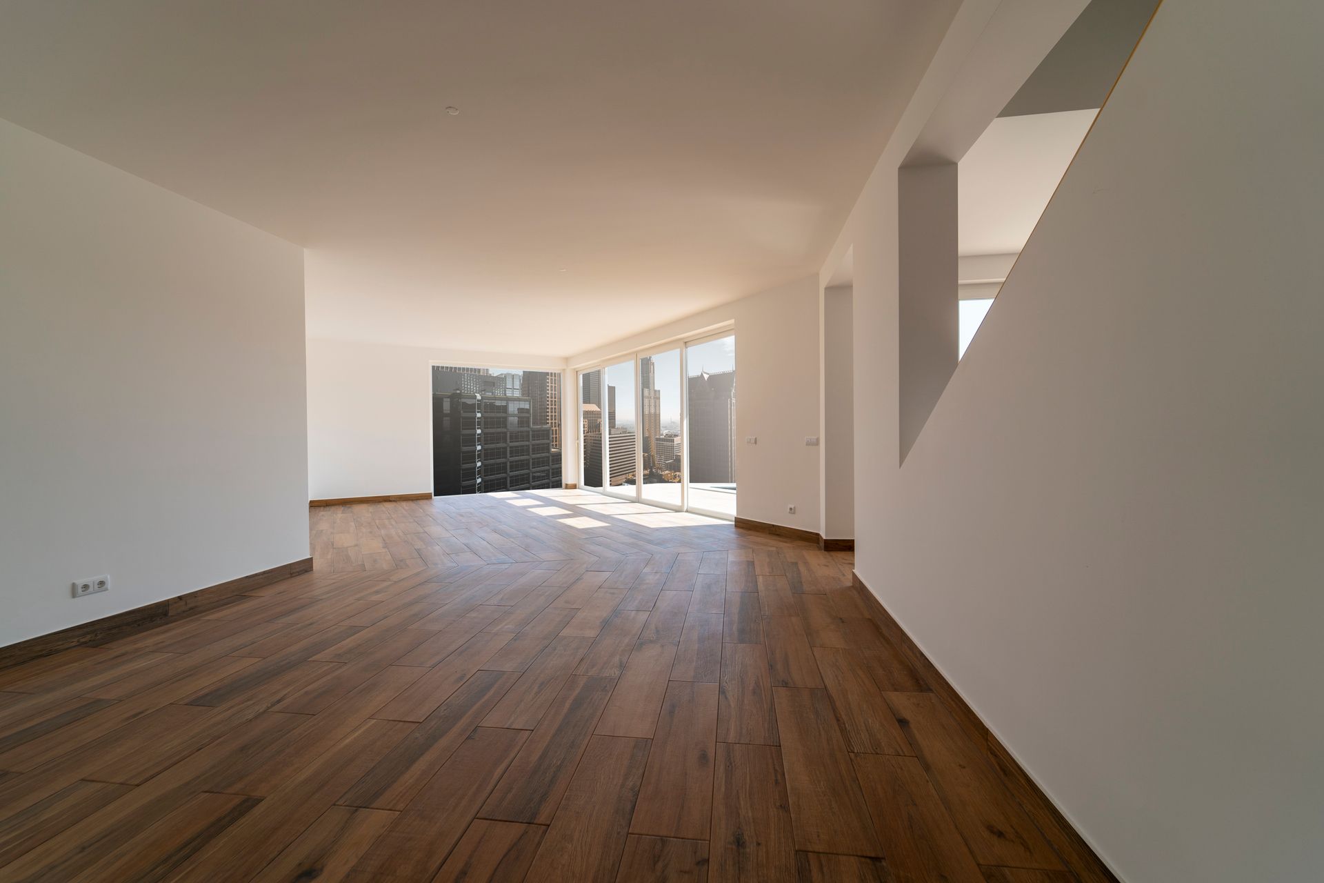Empty sunlit apartment room with wooden floors, white walls, and large windows looking out to a balcony.