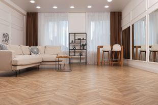 Bright modern living room with beige sofa, wooden herringbone floor, shelves, dining table, and large windows