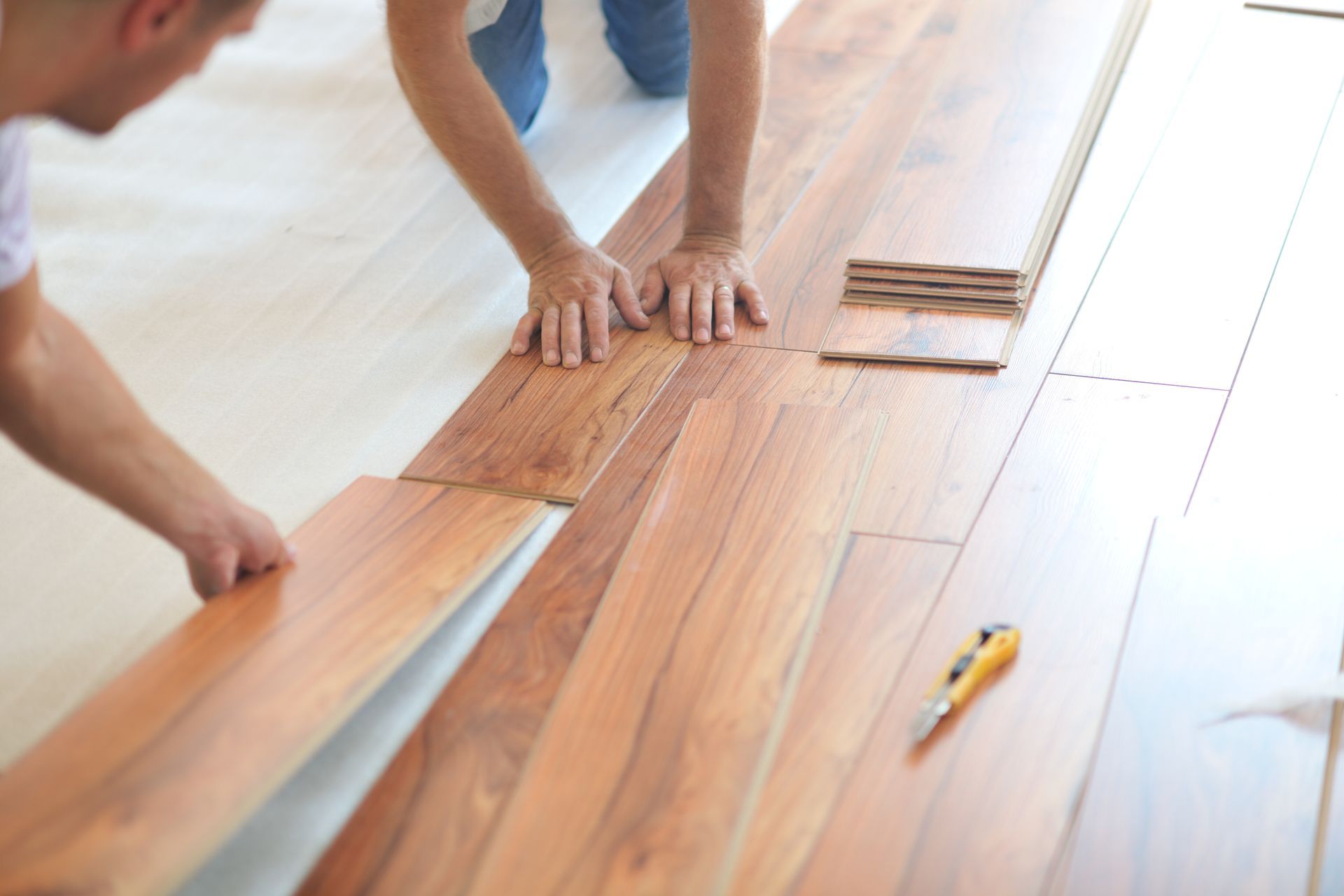 Two people installing wooden floor planks with tools on a bright room floor