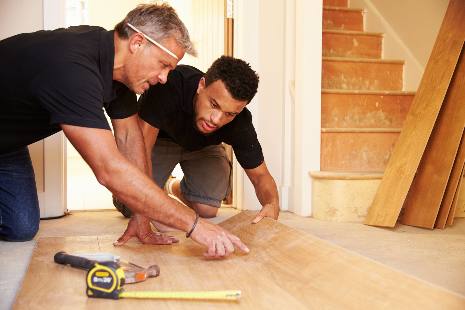 Two men kneeling on a wood floor, measuring and fitting planks during home renovation.