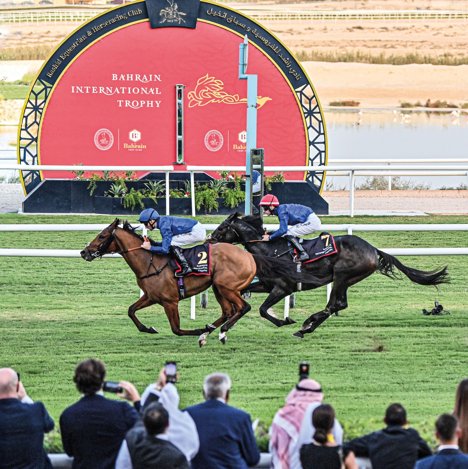 Two horses are racing in front of a bahrain international trophy sign