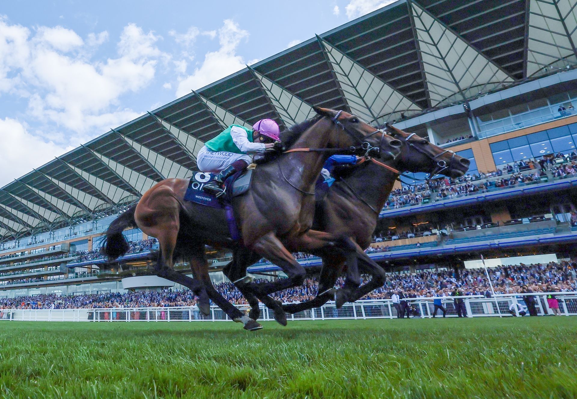 Two horses are racing on a race track in front of a crowd.