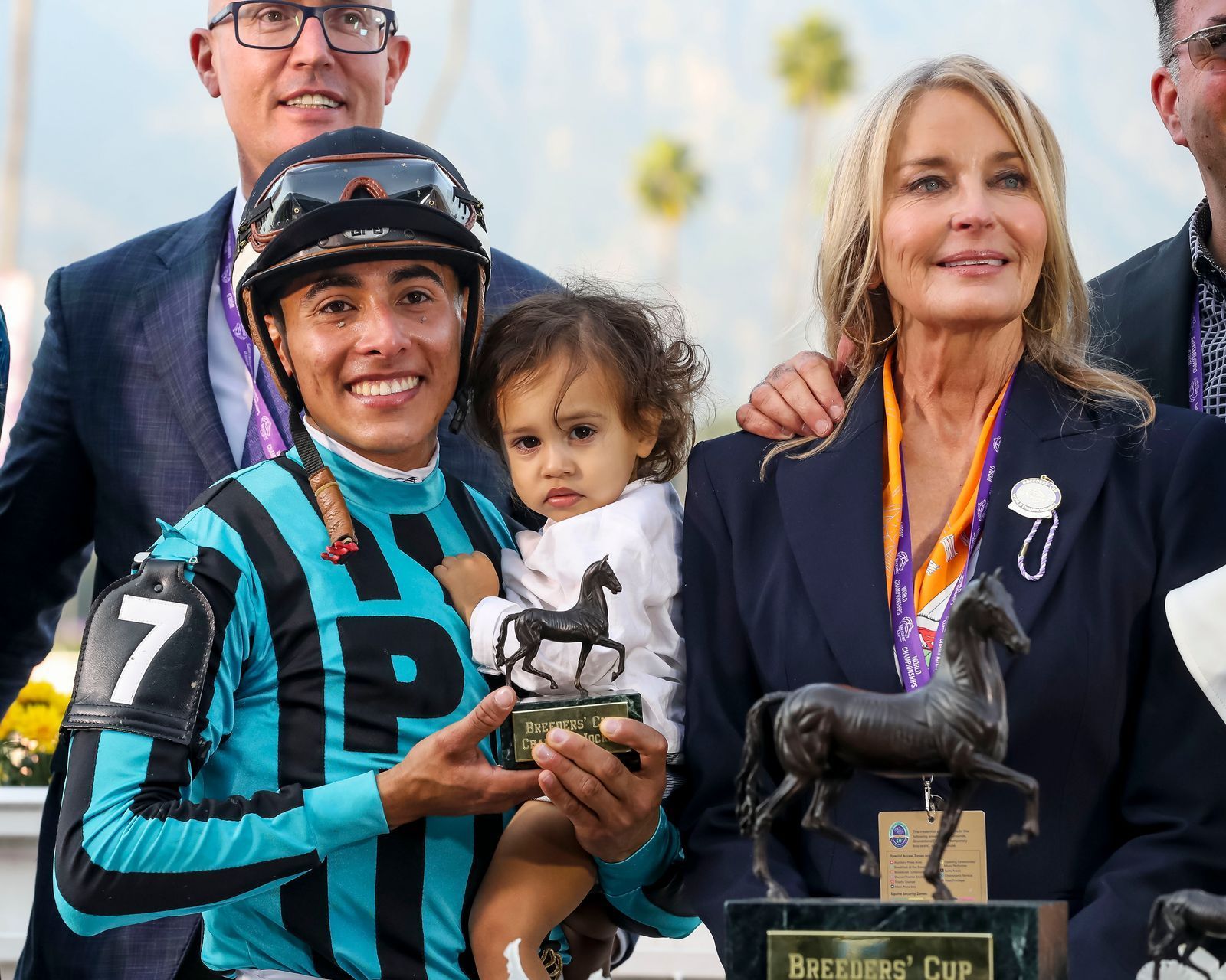 A jockey is holding a baby and holding a trophy.