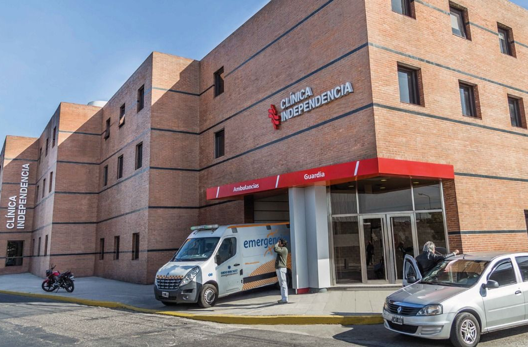 Entrada de hospital con ambulancia y personal. Edificio de ladrillo, detalles en rojo, día soleado.