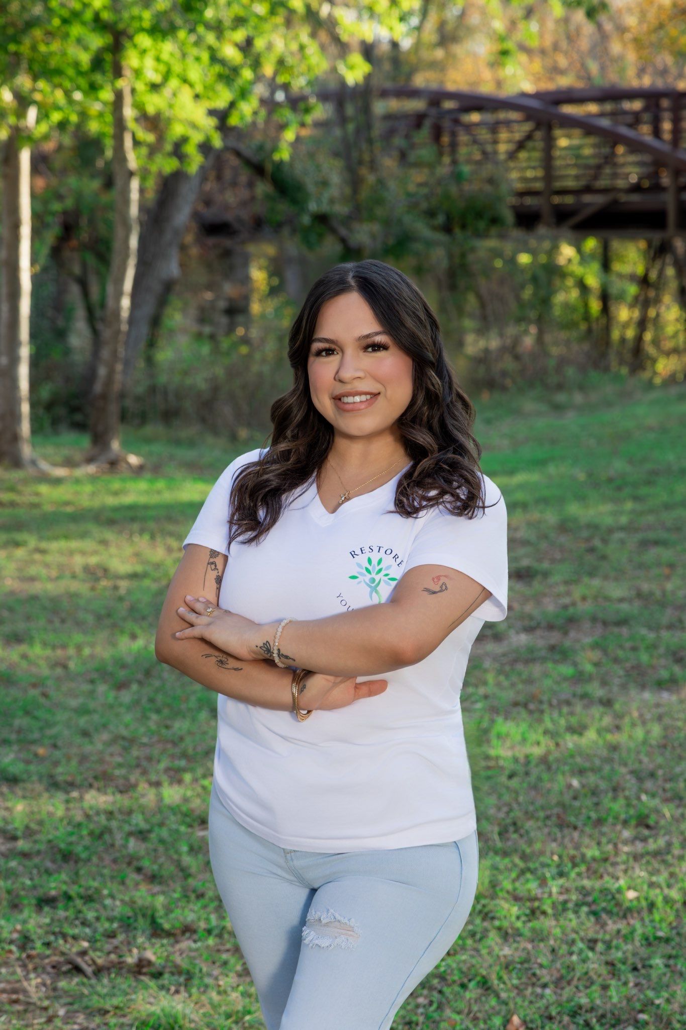 Woman in white shirt and ripped jeans smiling outdoors in a wooded area.