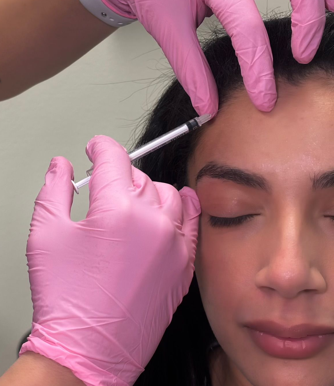 Person receiving facial filler injections, face close-up, pink gloves, syringes.