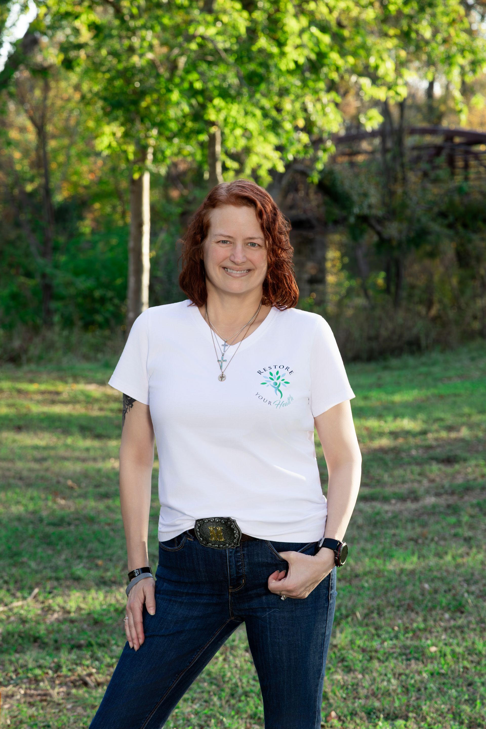 Woman with red hair in white top and jeans, hands in pockets, standing outdoors.