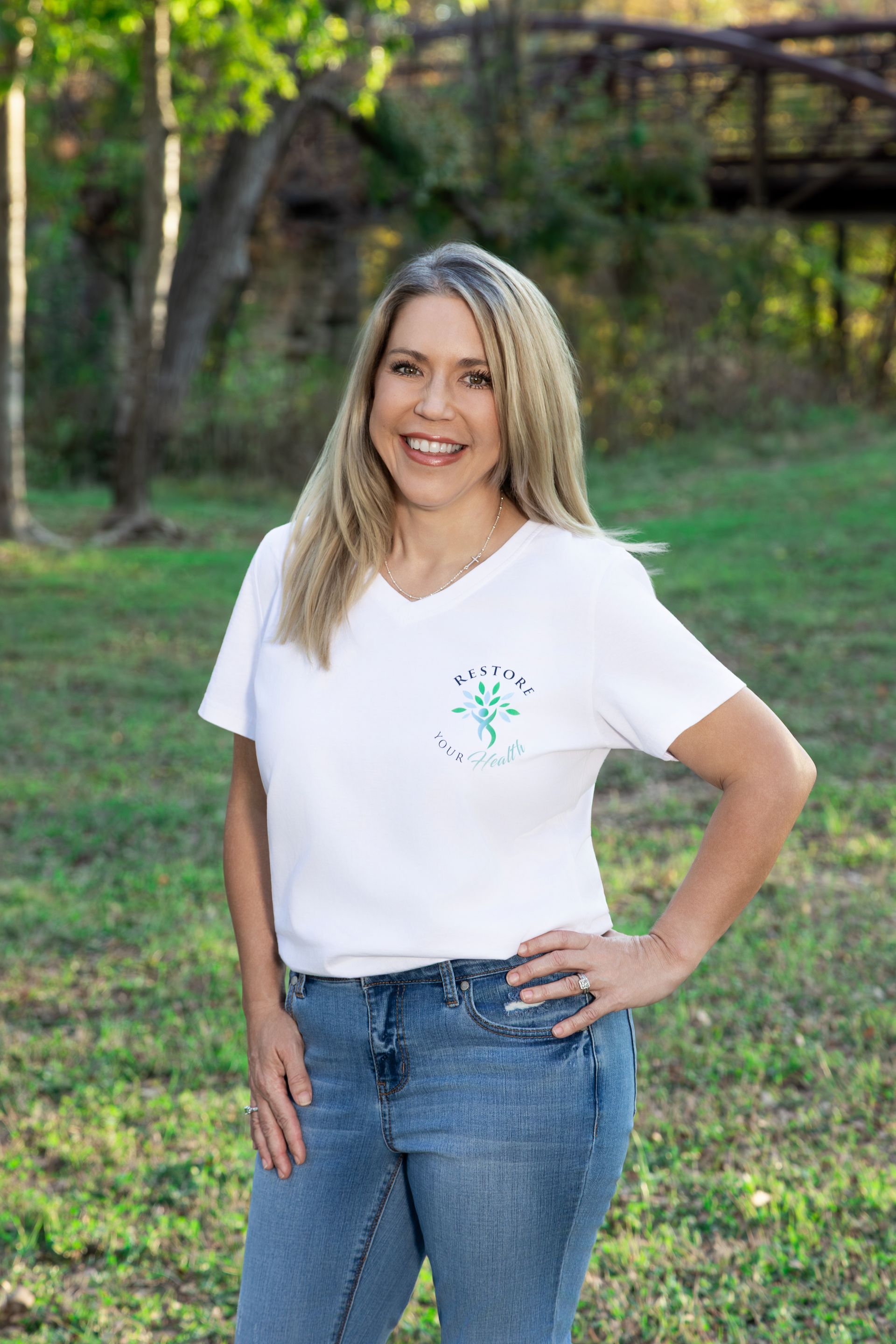 Blonde woman in white top and jeans smiles in a park, hands in pockets.
