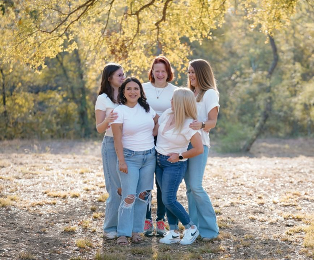 Five women in white shirts and jeans stand together outdoors under a tree, smiling.