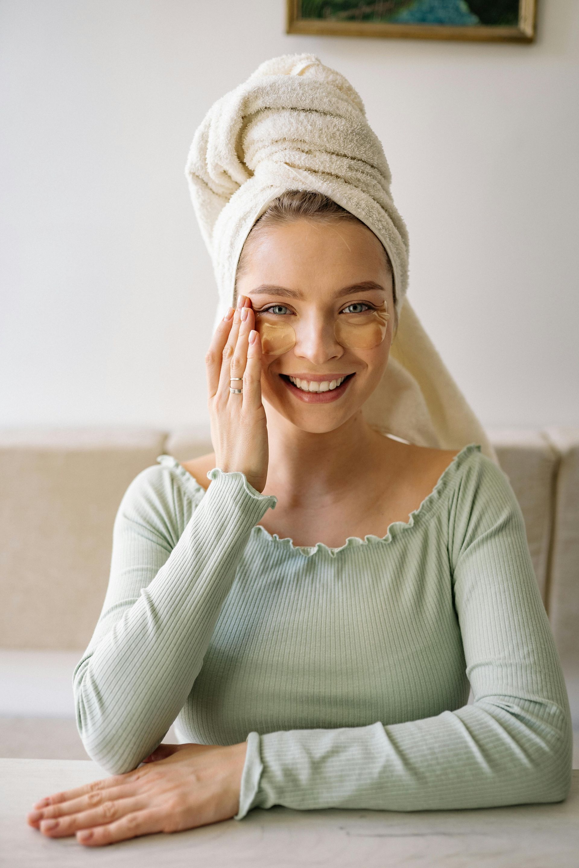 Woman with a towel on her head and under-eye patches smiling, sitting indoors.