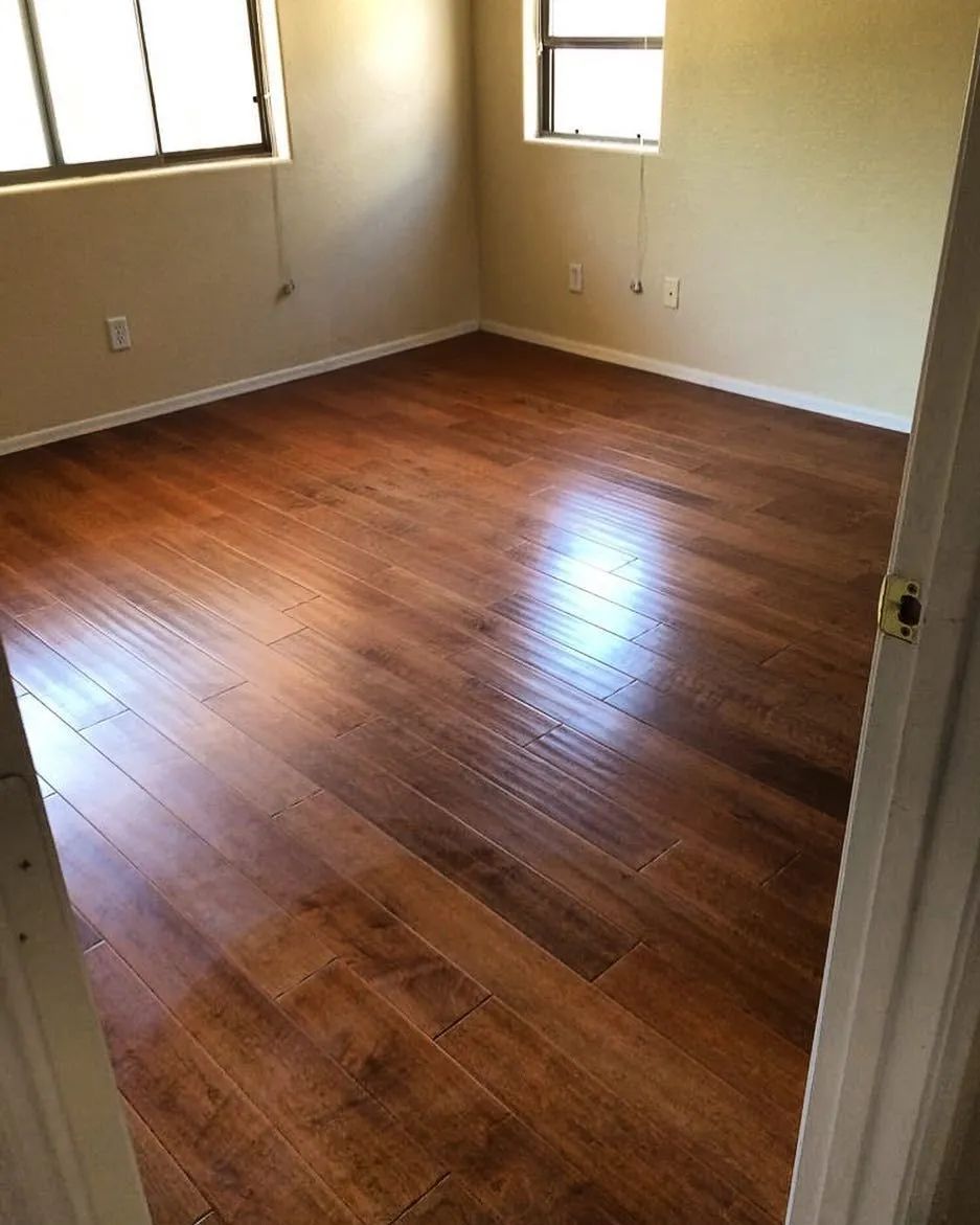 Empty room with glossy, dark wood floors and two windows. Beige walls, white doorframe.