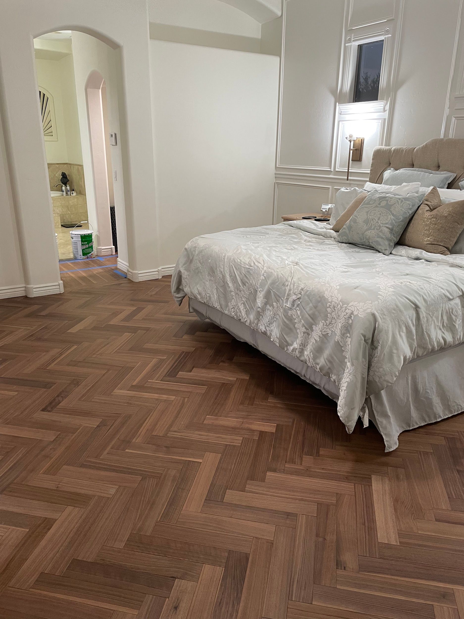 A bedroom with wood herringbone flooring, a bed with light-colored bedding and decorative pillows, and an open doorway.