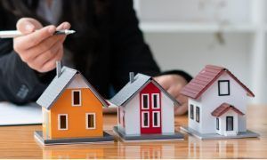 Person in a suit pointing at three miniature houses of different colors on a table.