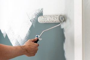 Hand painting a wall with a white paint roller, covering up a blue-gray wall.
