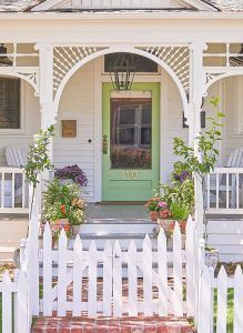 White cottage entrance with arched porch, green door, picket fence, and potted flowers.