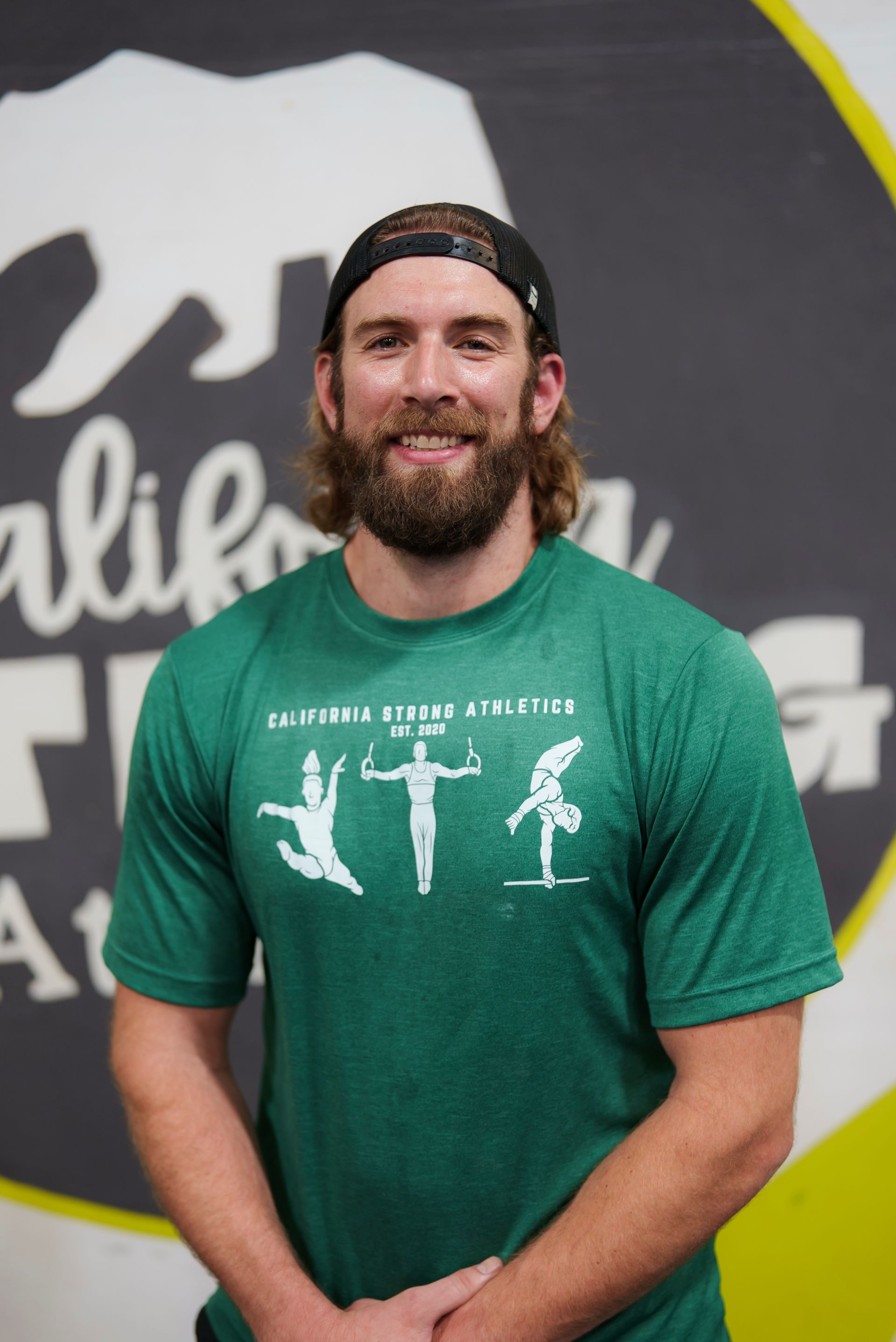 Man with beard and cap wearing green shirt stands in gym.