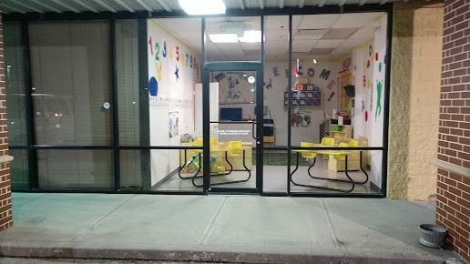 Exterior view of a brightly decorated children's classroom through glass windows, with yellow tables and colorful artwork.